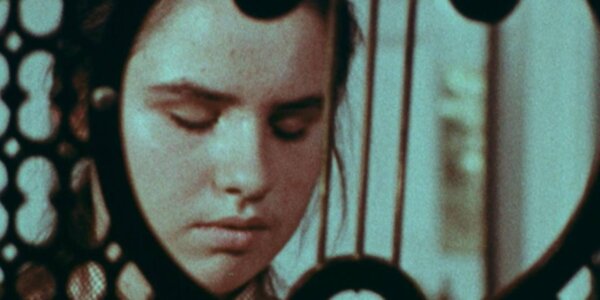 A still image taken from a grainy colour film, showing a close-up of a girl's face, her eyes closed. Slightly obscuring her face in the foreground is an empty sheet music stand.