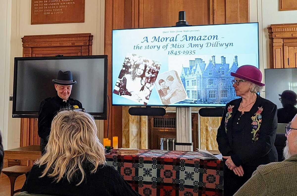 Dau berson yn rhoi cyflwyniad i gynulleidfa yn ystod digwyddiad 'Queer Tales from Wales' yn Ystafell y Cyngor yn Llyfrgell Genedlaethol Cymru