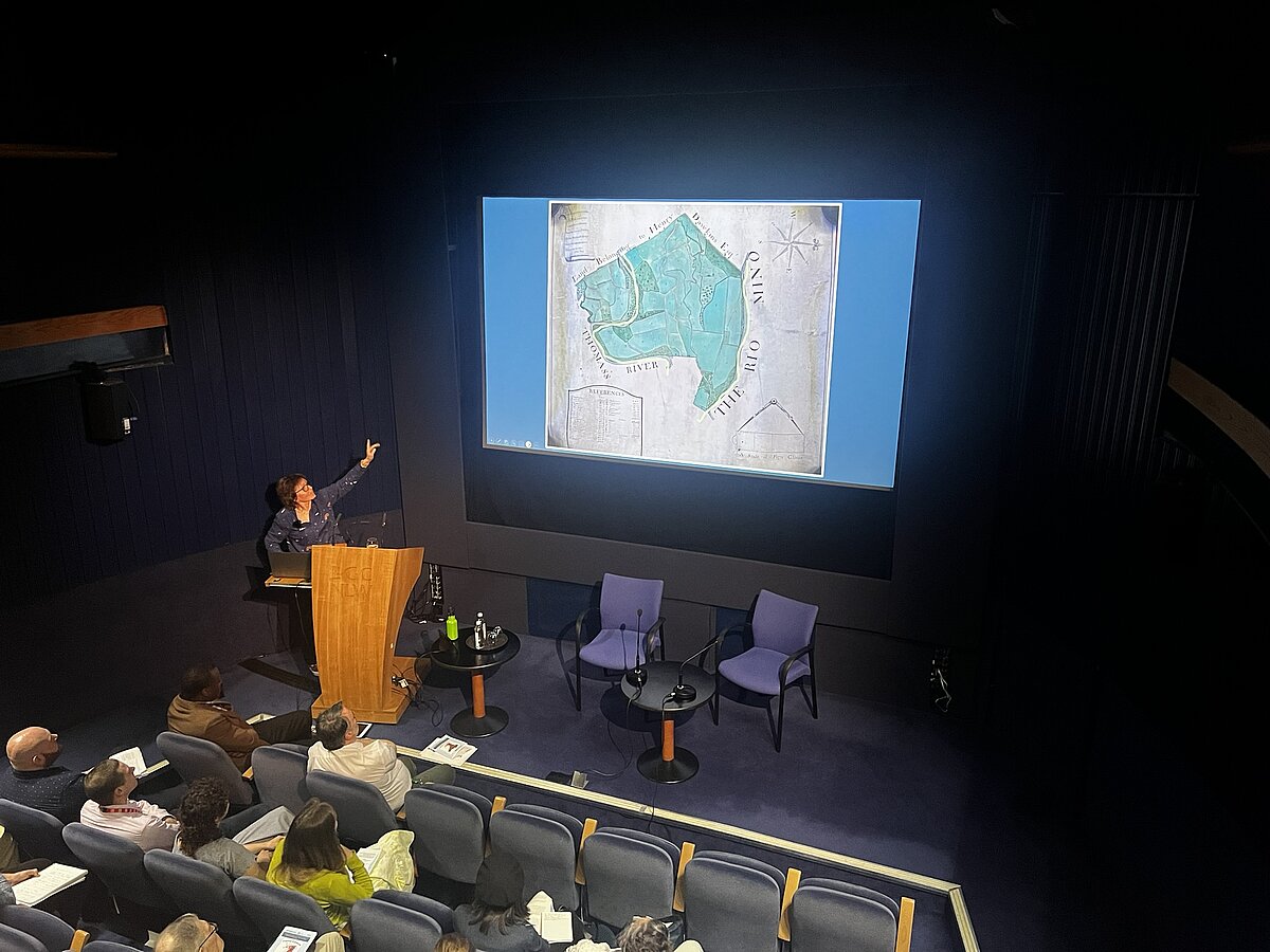 Mae Marian Gwyn yn ystumio at sgrin sy'n dangos map llawysgrif o blanhigfa
