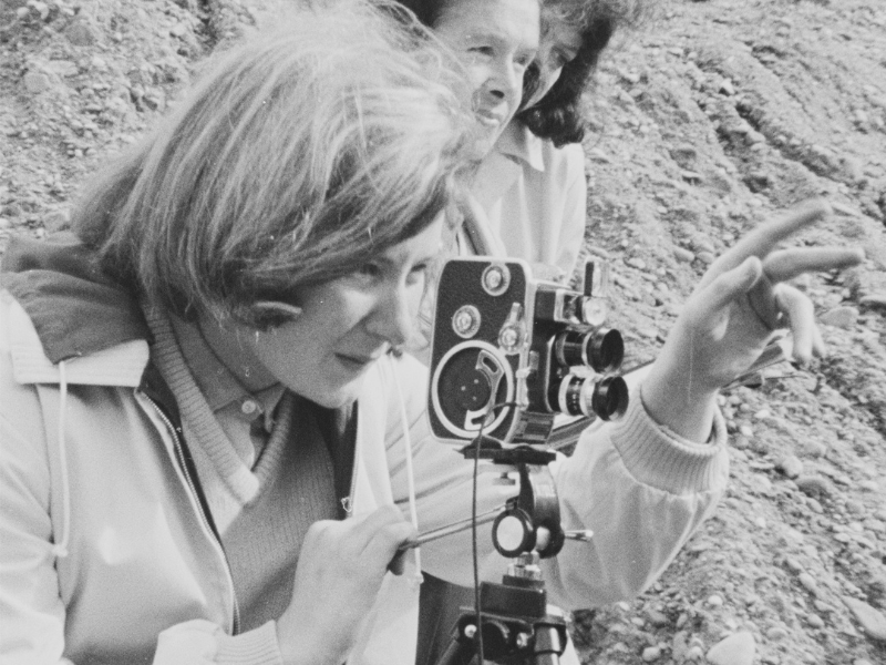A black and white still image from a news report on the filming of amateur film 'From Wrexham With Love', which shows a close up of a woman looking through an 8mm camera lens, and pointing out of frame.