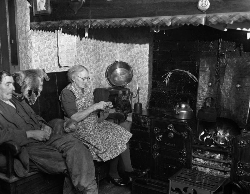 Llun du a gwyn o dau berson yn eistedd wrth aelwyd mewn ffermdy gyda canhwyllau brwyn yn sychu uwchben y tan